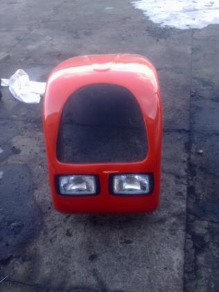 Czerwona, błyszcząca obudowa kabiny małego pojazdu z dwoma reflektorami leży na szarym, betonowym podłożu.