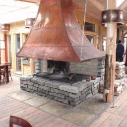 Rustykalny kominek z miedzianym okapem i kamienną obudową, z rusztem i akcesoriami do grillowania, umieszczony na drewnianym tarasie w zadaszonej przestrzeni, z widocznymi elementami oświetlenia...