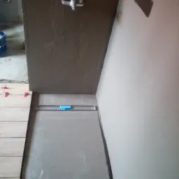 Montaż płytek w łazience: widoczny fragment ściany z nałożonym klejem, poziomica, ułożone płytki podłogowe z systemem poziomowania, bateria prysznicowa na ścianie.