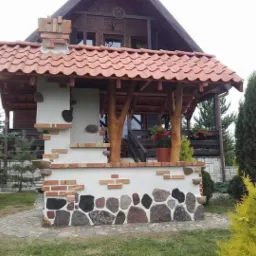 Murowany grill ogrodowy z cegły i kamienia, z drewnianym zadaszeniem krytym dachówką, na tle drewnianego domu.