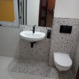 Umywalka i toaleta w wykończonej łazience z mozaikową okładziną ścienną i podłogową w odcieniach bieli i szarości, czarna armatura, widoczne okno z roletą i odbicie fotografa w lustrze.