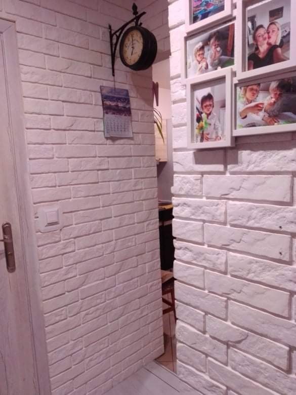 Ściana wykończona białą cegłą dekoracyjną z zawieszonym zegarem i kalendarzem oraz ramkami ze zdjęciami rodzinnymi.