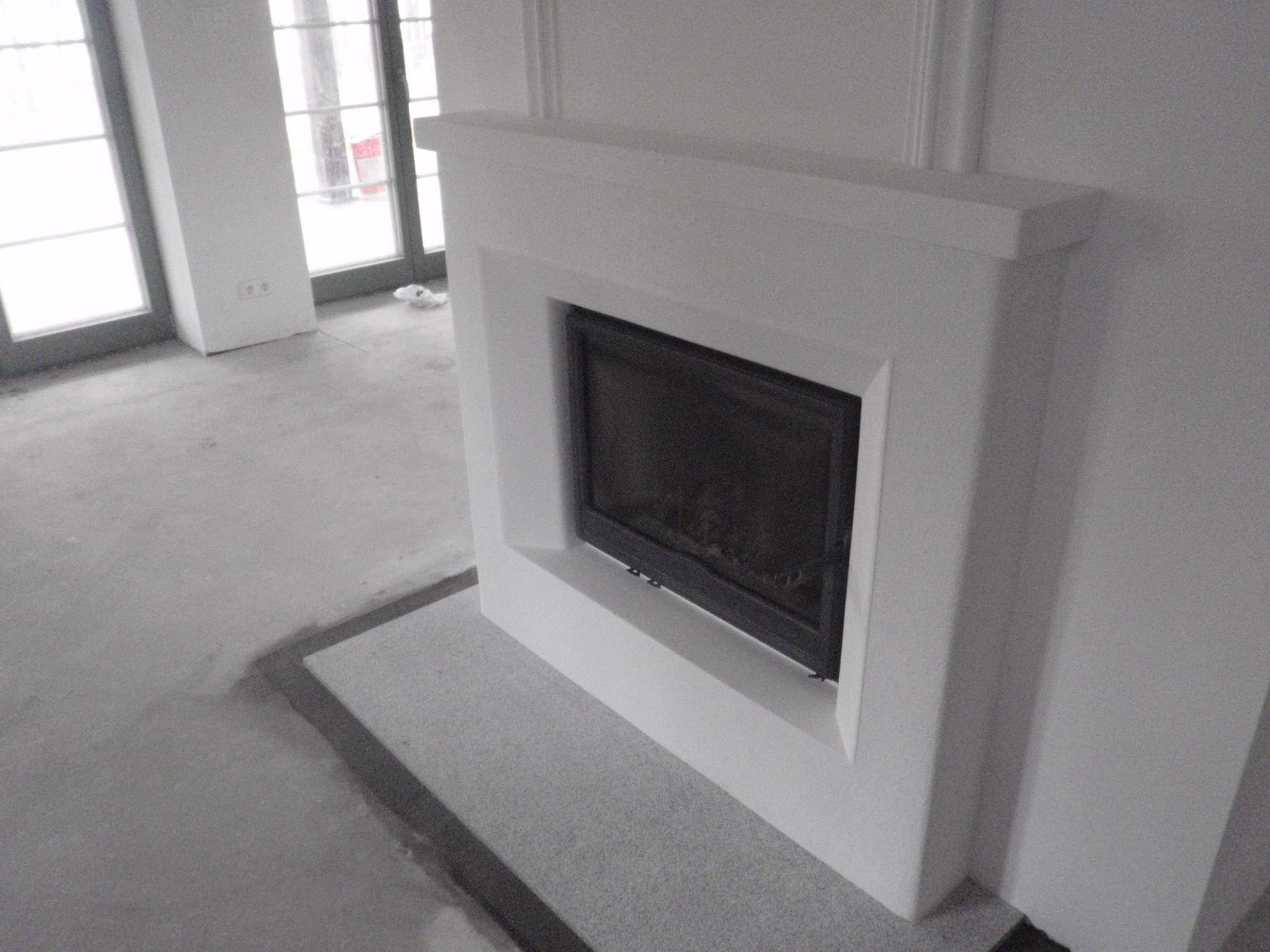 Biały, minimalistyczny kominek z czarnym wkładem, osadzony na szarym, kamiennym podłożu, widok z boku, surowy stan wykończenia podłogi.