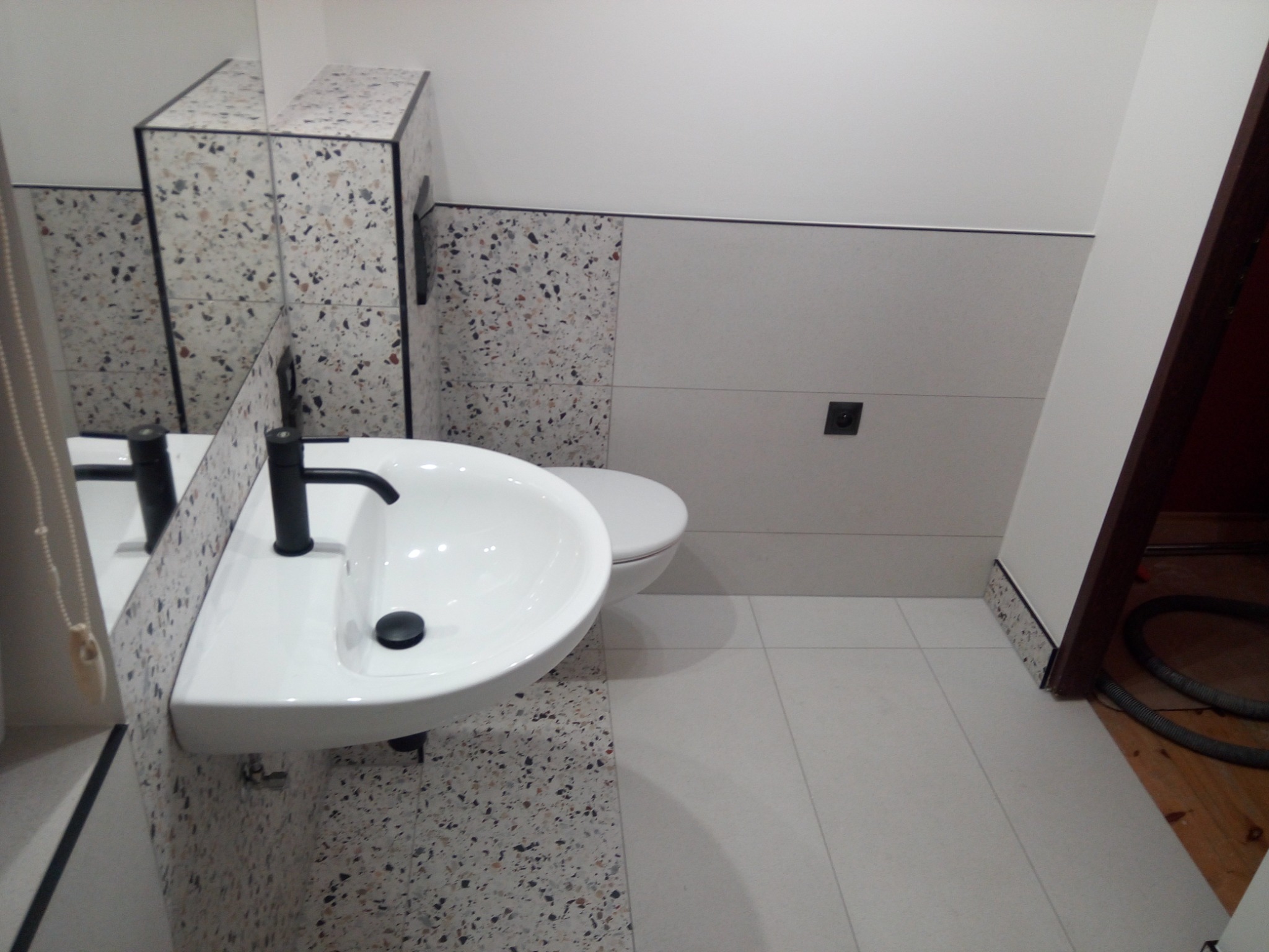 Nowoczesna łazienka z umywalką nablatową, toaletą i płytkami imitującymi lastryko na ścianach i podłodze, widoczny fragment odkurzacza przy progu drzwi.