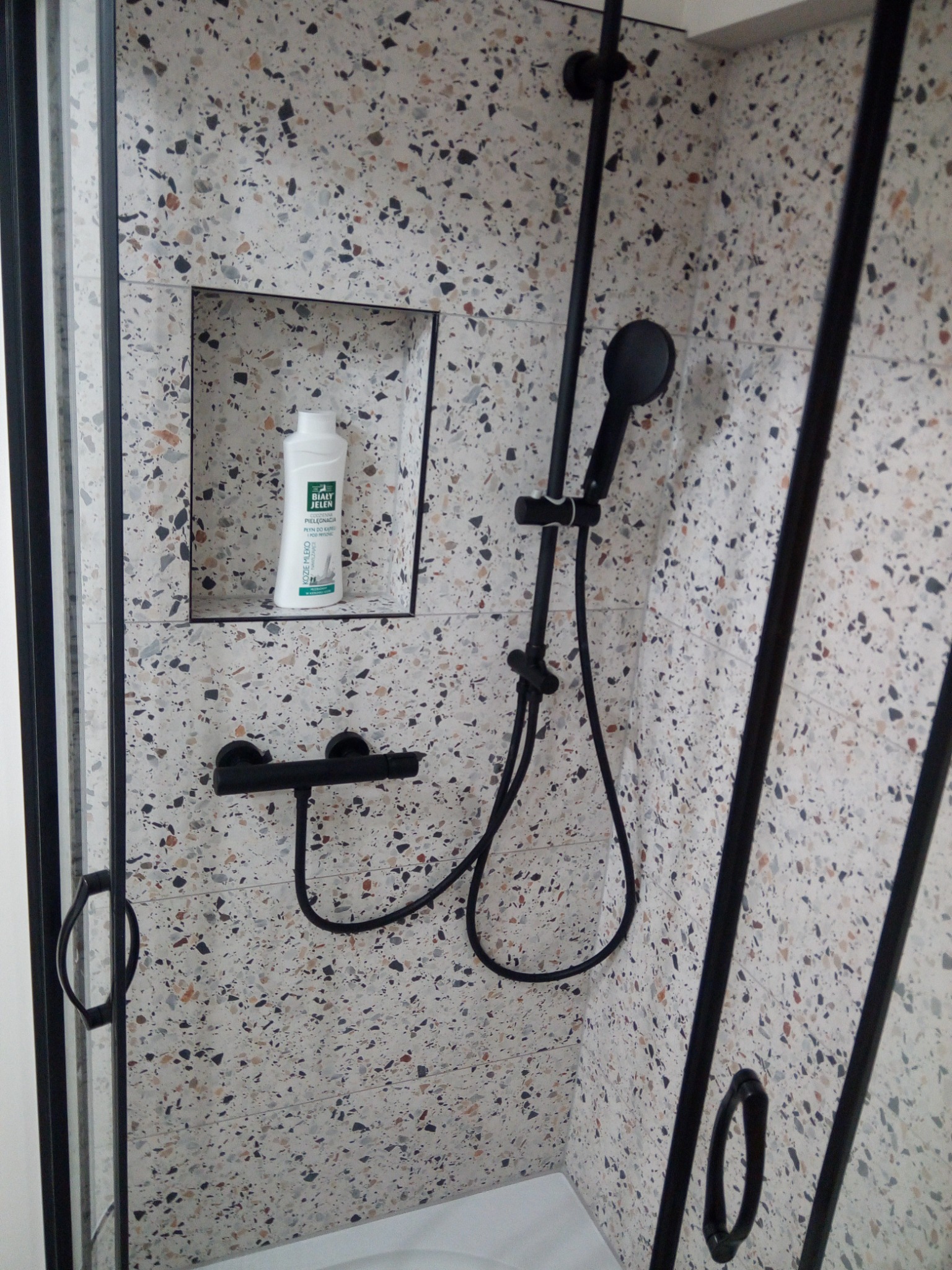 Kabina prysznicowa z czarną armaturą i ścianami wyłożonymi płytkami imitującymi lastryko, z wnęką na kosmetyki, w której stoi butelka płynu do kąpieli.