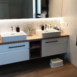 Nowoczesna łazienka z dwoma umywalkami nablatowymi, drewnianym blatem, białymi szafkami z czarnymi uchwytami, dużym lustrem z podświetleniem LED i teksturowanymi płytkami na ścianie, uzupełniona...