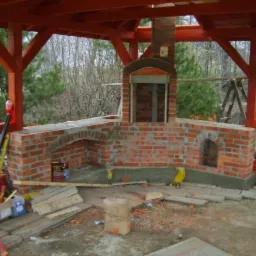 Murowana konstrukcja z cegły, prawdopodobnie piec lub kominek zewnętrzny, w trakcie budowy pod drewnianą konstrukcją z czerwonymi belkami. Widoczne materiały budowlane i narzędzia. Otoczenie...