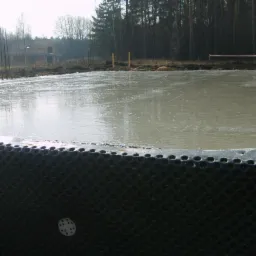 Wylany fundament pod dom jednorodzinny z widoczną folią kubełkową i fragmentem budynku w tle, w pochmurny dzień.