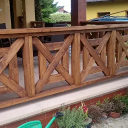Drewniana balustrada balkonowa z krzyżowym wzorem, zabezpieczona lakierem, z widocznymi detalami słoj&oacute;w drewna, na tle elewacji domu i zieleni.