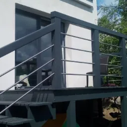 Zewnętrzne schody z szarego drewna i metalową balustradą prowadzące na taras przy białym budynku mieszkalnym, widoczne buty i krzesło.