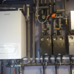 Nowoczesny, biały piec gazowy marki Brotje zamontowany na ścianie, z widocznymi rurami i elementami instalacji grzewczej, w tym czarne regulatory i izolowane rury.