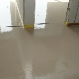 Świeżo wylana, mokra posadzka w jasnym odcieniu beżu, z widocznymi elementami przygotowania podłoża przy ścianach i instalacją grzewczą w tle.