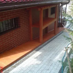 Zadaszone wejście do budynku z czerwonej cegły, z drewnianą konstrukcją wspartą na słupach, widoczne panele podłogowe i czerwony dach z rynnami.