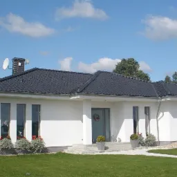 Nowoczesny dom jednorodzinny z ciemną dachówką, anteną satelitarną i zadbanym trawnikiem w słoneczny dzień.