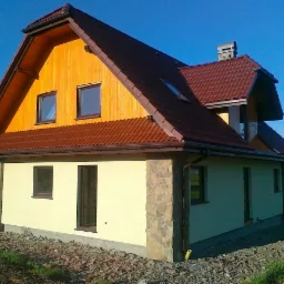 Dwukondygnacyjny dom z jasną elewacją, drewnianą górną częścią szczytu i brązową dachówką, z widocznym kominem i oknem dachowym, otoczony kamieniami i trawą.