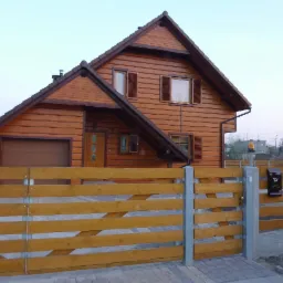 Gotowy, dwukondygnacyjny dom z bali z drewnianym ogrodzeniem, skrzynką pocztową i automatyczną bramą wjazdową, widok z zewnątrz, w tle drzewa i zabudowania.