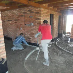 Wnętrze w trakcie budowy z ceglanymi ścianami, widoczne szare rury na podłodze i dwóch robotników w trakcie prac wykończeniowych, jeden w czerwonej koszulce oparty o maszynę, drugi w niebieskiej...