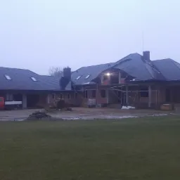 Dom jednorodzinny w trakcie budowy, z widoczną konstrukcją dachu pokrytą ciemną dachówką, rusztowania, cegły i czerwony samochód dostawczy na posesji w pochmurny dzień.