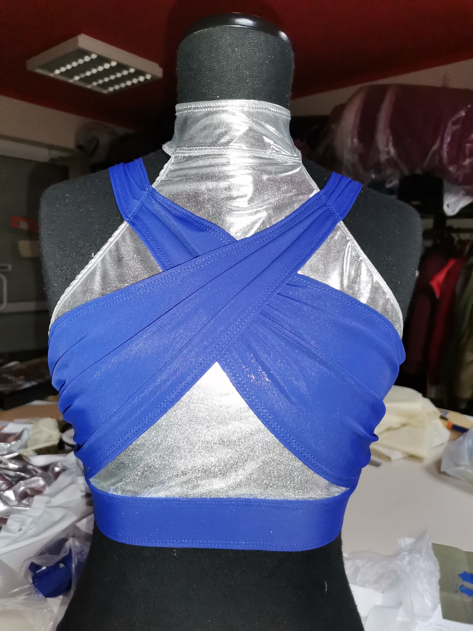 Niebieski top z krzyżującymi się ramiączkami na srebrnym manekinie krawieckim z widocznymi szwami i detalami wykończenia, uchwycony w szwalni z widocznym oświetleniem sufitowym i fragmentami innych...