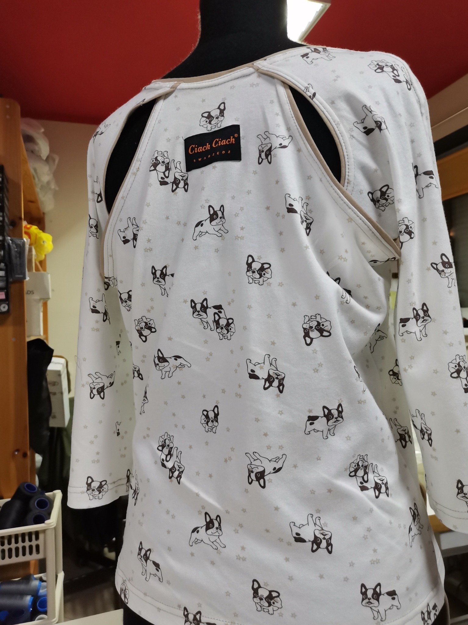 Biała bluzka z długim rękawem, z rozcięciami na ramionach i wzorem w buldogi francuskie, prezentowana na manekinie, z widoczną metką producenta.