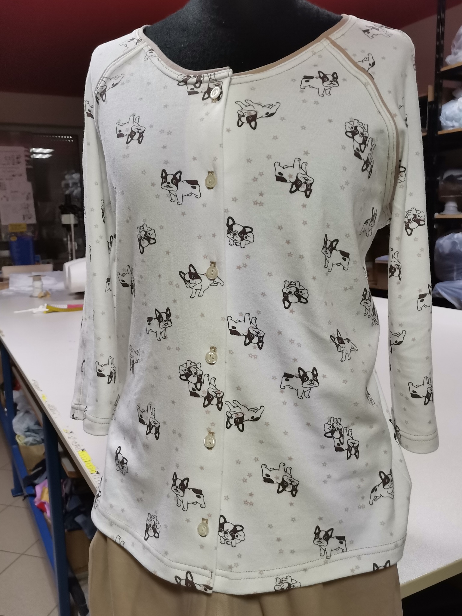 Jasna bluzka z długim rękawem, zapinana na guziki, w dziecięcy wzór z buldogami francuskimi i gwiazdkami, prezentowana na manekinie w szwalni.