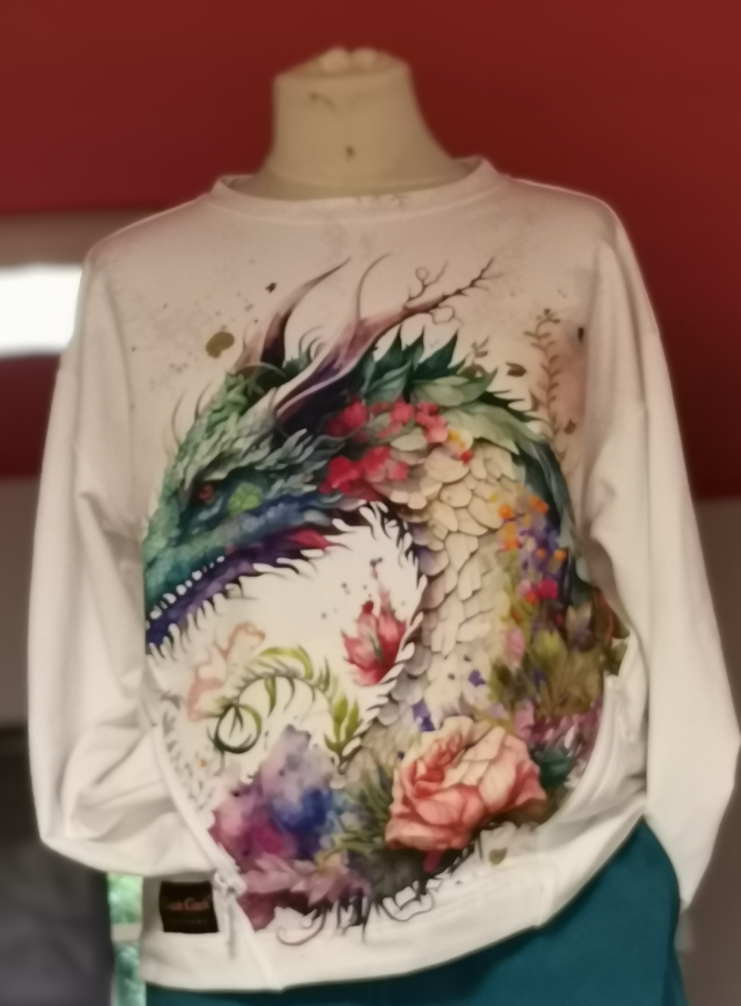 Biała bluza z długim rękawem prezentowana na manekinie, z dużym, barwnym nadrukiem smoka otoczonego kwiatami w stylu akwareli.