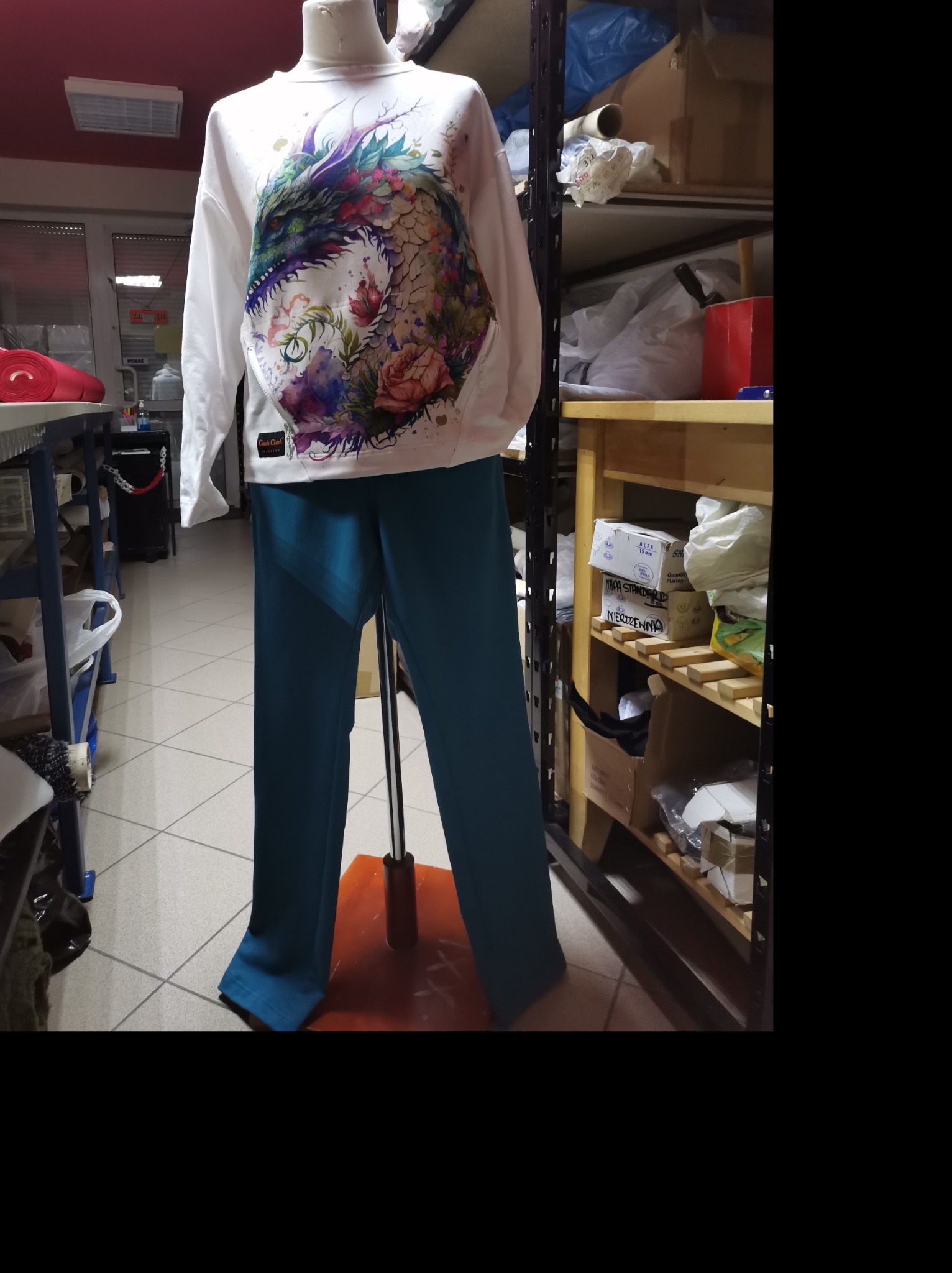 Manekin prezentuje białą bluzę z kolorowym nadrukiem smoka i niebieskie spodnie w pomieszczeniu z regałami