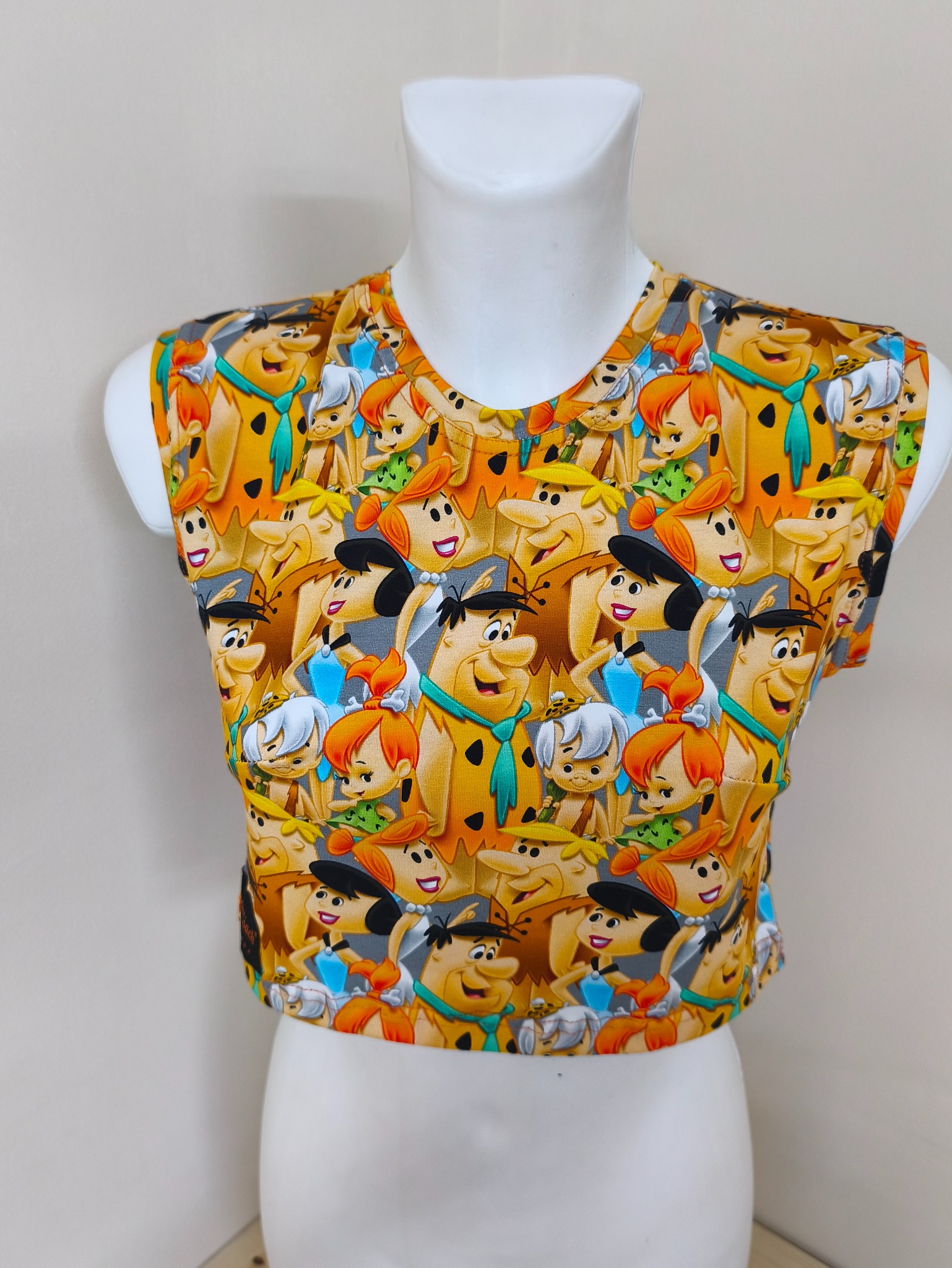 Damska bluzka bez rękawów z kolorowym wzorem przedstawiającym postacie z kreskówki 'Flintstonowie', prezentowana na białym manekinie.