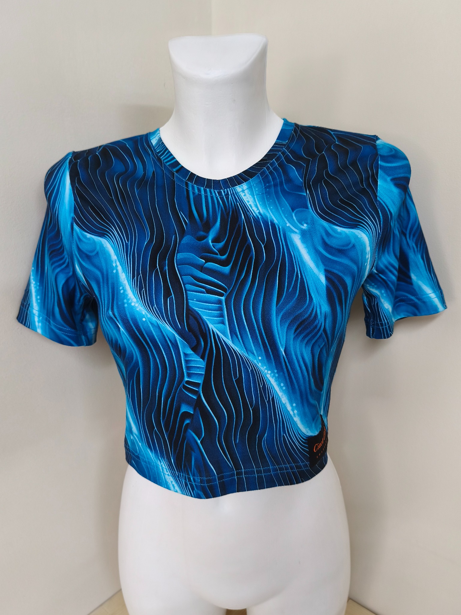 Krótka, dopasowana bluzka z graficznym, niebieskim wzorem na manekinie. Widoczny krój i detale wykończenia. Bluzka prezentowana na neutralnym tle.