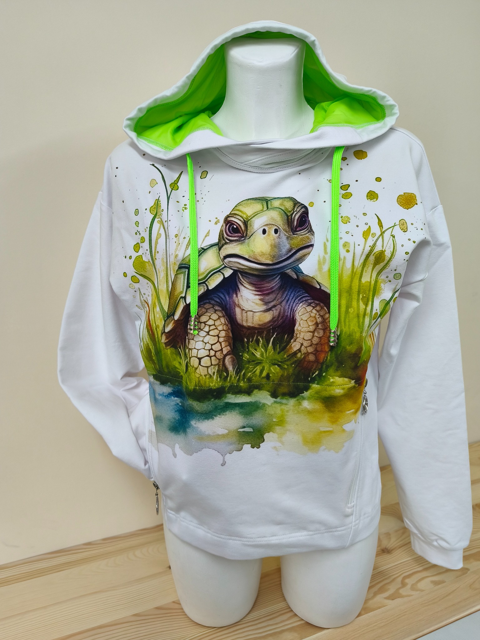 Biała bluza z kapturem i nadrukiem żółwia na manekinie. Kaptur wykończony jaskrawozieloną podszewką i sznurkami. Widoczny zamek błyskawiczny z boku.