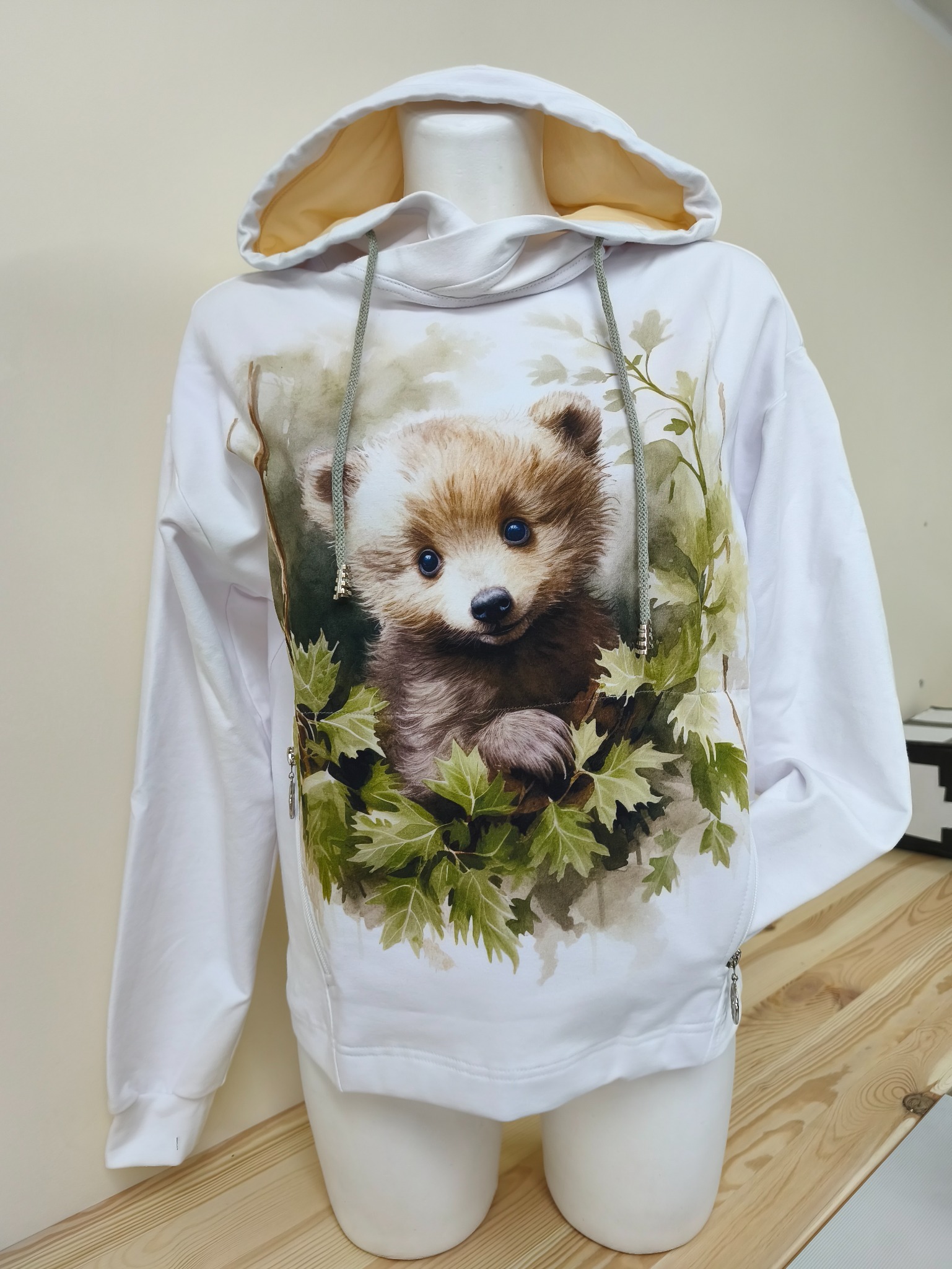 Biała bluza z kapturem, z nadrukiem uroczego misia w liściach, prezentowana na manekinie w Swarzędzu. Bluza z zamkami po bokach i sznurkami w kapturze.