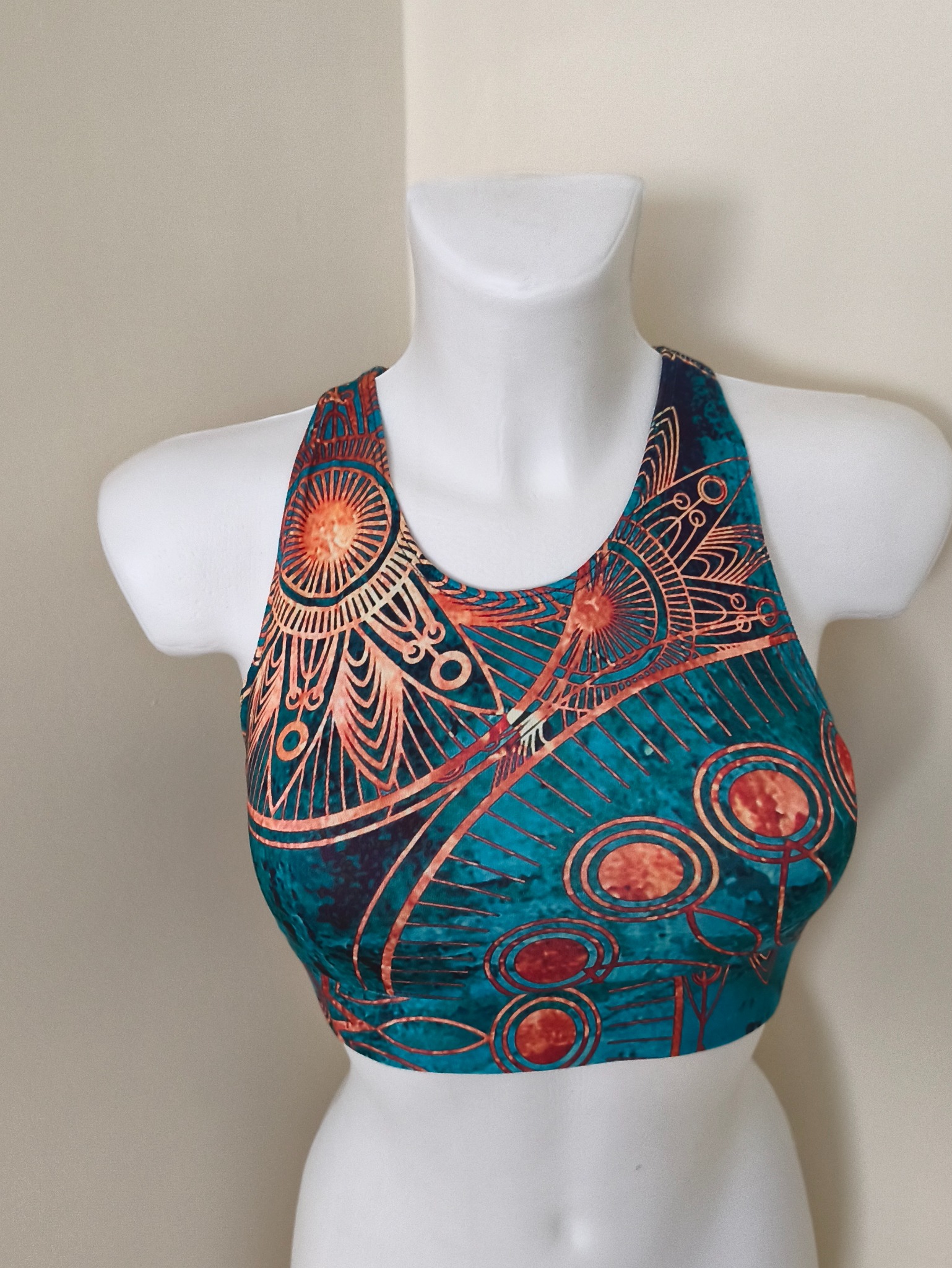 Turkusowy top sportowy z pomarańczowymi, abstrakcyjnymi wzorami na białym manekinie, na tle jasnej ściany. Wyraźny detal wzoru i krój.