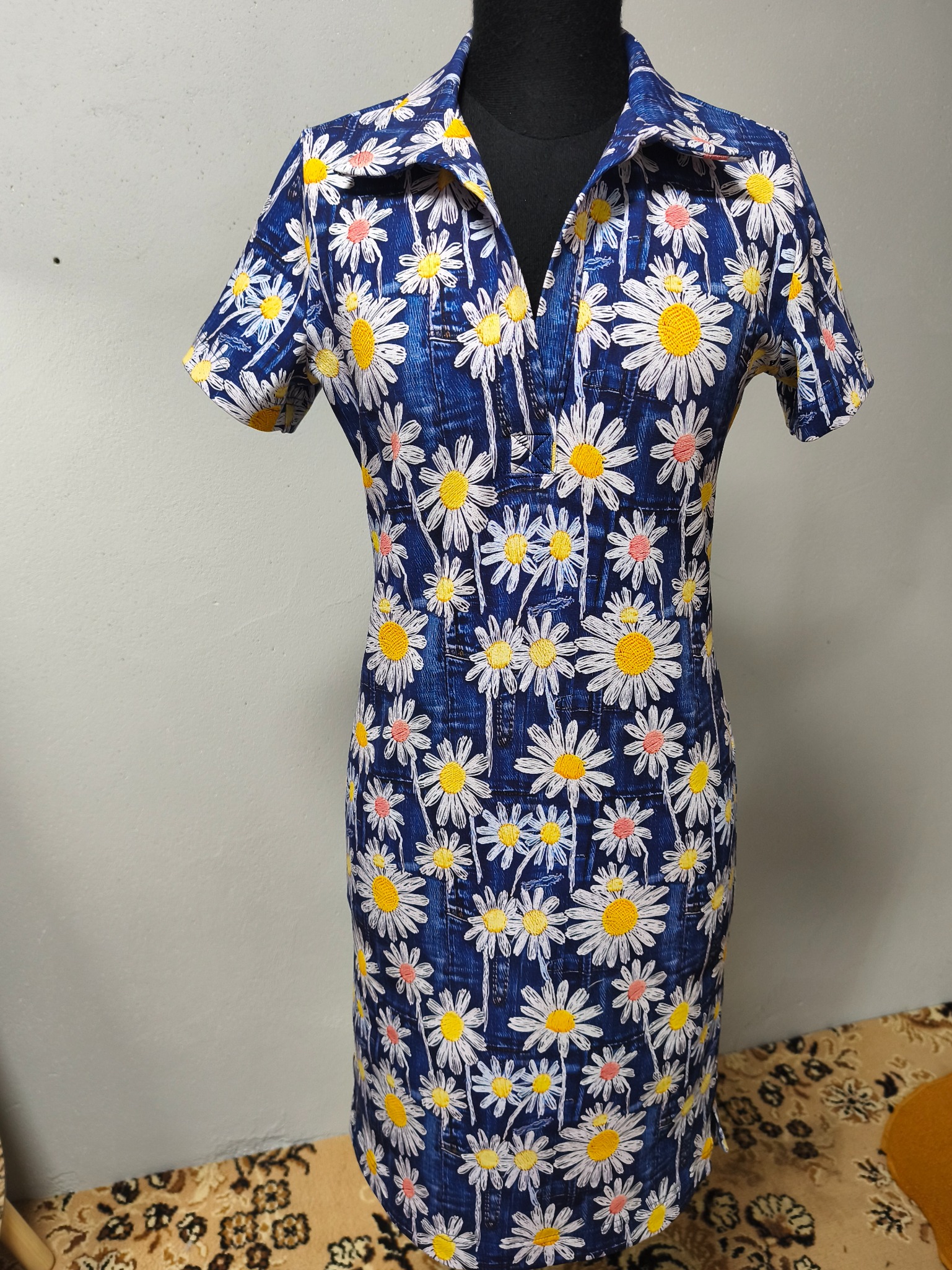 Sukienka w kwiaty na manekinie, krótki rękaw, styl retro z lat 70., niebieska tkanina w białe i żółte stokrotki. Widoczne detale wykończenia i krój.
