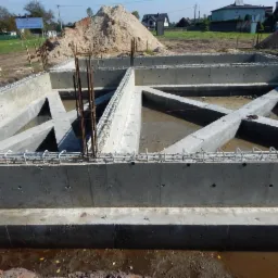 Zalane fundamenty domu z widocznym zbrojeniem i fragmentami szalunków. W tle hałda piasku i zabudowa jednorodzinna w słoneczny dzień.