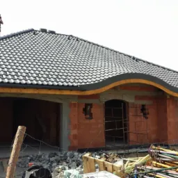 Dom w stanie surowym otwartym z ciemną dachówką ceramiczną. Widoczny mur z czerwonej cegły i drewniane elementy konstrukcyjne. Na dachu stoi osoba.