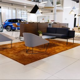 WITAN Centrum Podłóg Sp. z o.o. - Nowoczesne wnętrze salonu z pomarańczowym dywanem, szarą sofą i krzesłami na tle jasnej podłogi i ekspozycji aut. Minimalistyczne lampy wiszące.