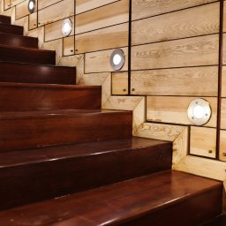 WITAN Centrum Podłóg Sp. z o.o. - Drewniane schody z ciemnego drewna, oświetlone punktowymi lampami w ścianie z jasnego drewna. Nietypowe wykończenie ścian z drewnianych elementów.