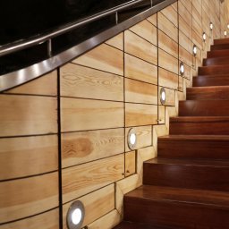 WITAN Centrum Podłóg Sp. z o.o. - Drewniane schody z oświetleniem punktowym i metalową barierką, prowadzące w górę, z widoczną strukturą drewna i ciemnym tłem.