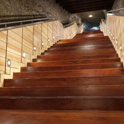 WITAN Centrum Podłóg Sp. z o.o. - Drewniane schody z oświetleniem punktowym wzdłuż ściany z jasnego drewna, prowadzące w górę do oświetlonego tunelu. Metalowa barierka po lewej stronie.