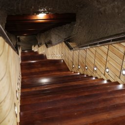 WITAN Centrum Podłóg Sp. z o.o. - Schody z ciemnego drewna z podświetleniem LED wzdłuż jasnych, drewnianych ścianek, prowadzące w dół w kamiennym tunelu z metalową poręczą.