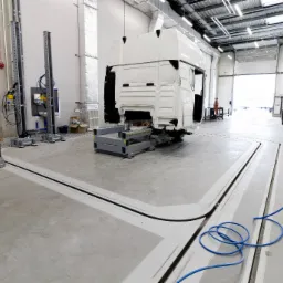 Wnętrze hali warsztatowej z kanałem naprawczym i podnośnikami, przygotowane do naprawy kabiny ciężarówki. Widoczne narzędzia i oświetlenie.