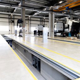 WITAN Centrum Podłóg Sp. z o.o. - Hala warsztatowa z kanałem naprawczym, betonową posadzką i żółtymi pasami. W tle widoczne naprawiane samochody ciężarowe oraz pracownicy serwisu.