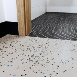 WITAN Centrum Podłóg Sp. z o.o. - Fragment podłogi z żywicy epoksydowej w neutralnym odcieniu z kolorowymi wtrąceniami, przechodzącej w podłogę z blachy ryflowanej w nowym pomieszczeniu.