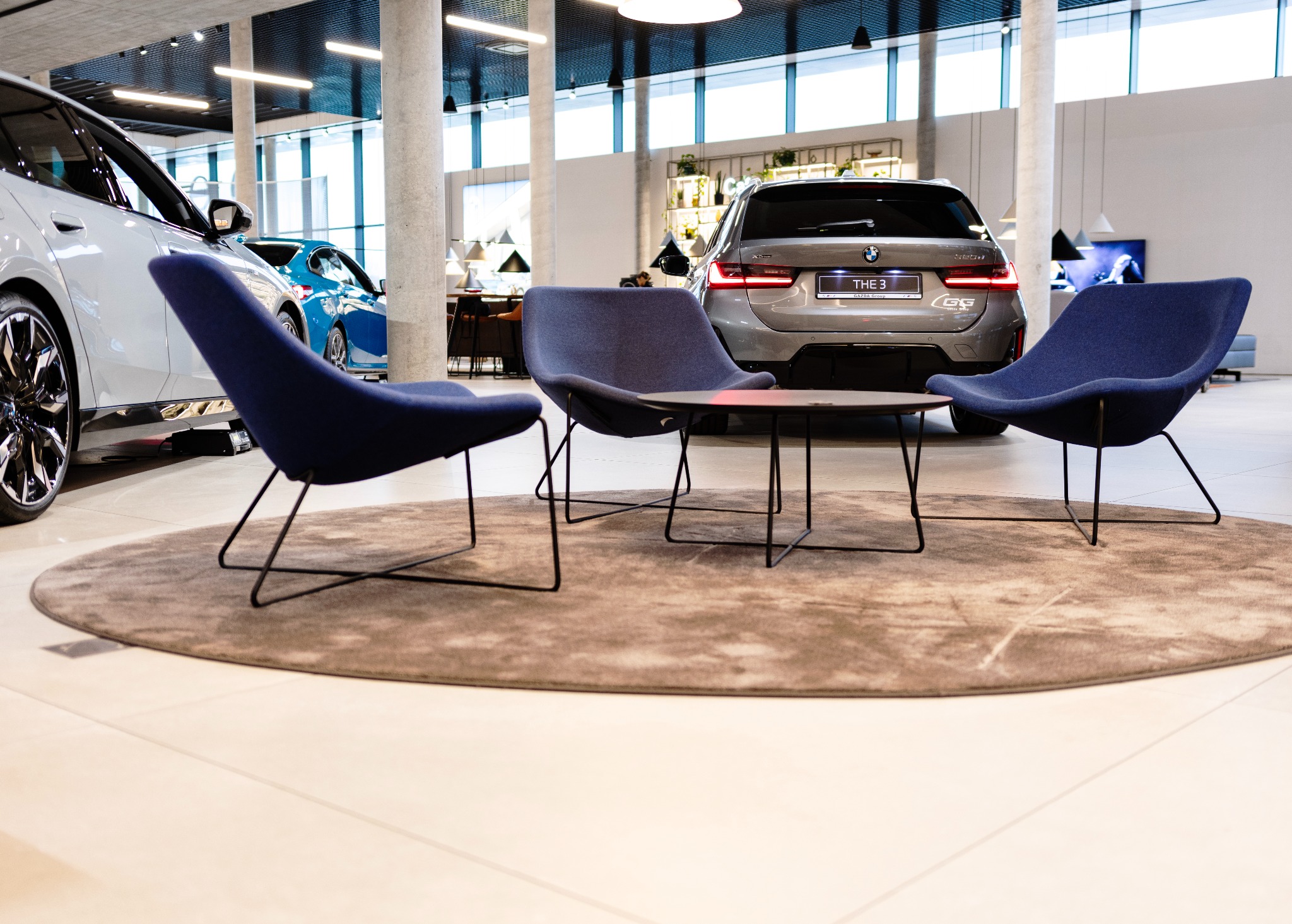 Jasna podłoga z okrągłym, beżowym dywanem, na którym stoją trzy granatowe krzesła i stolik. W tle samochody w salonie sprzedaży.