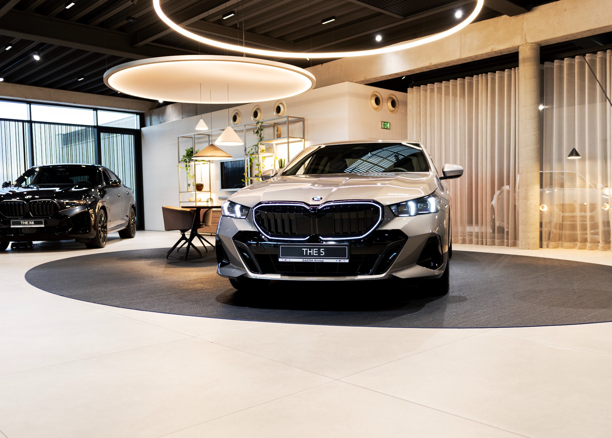 Salon samochodowy z beżowym BMW THE 5 na okrągłej wykładzinie. W tle czarny BMW X6, designerskie lampy i meble. Nowoczesne oświetlenie i minimalistyczny wystrój.
