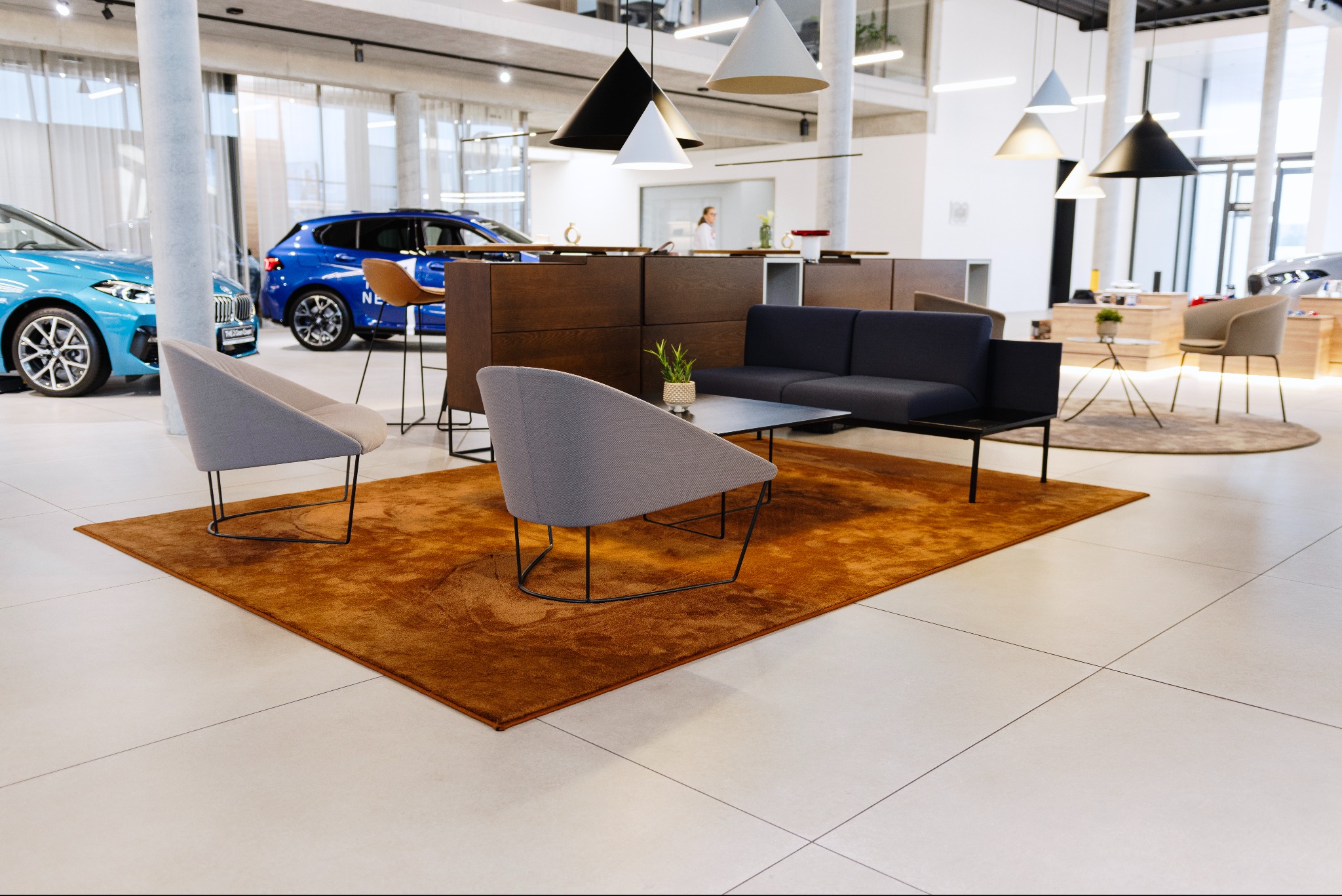 Nowoczesne wnętrze salonu z pomarańczowym dywanem, szarą sofą i krzesłami na tle jasnej podłogi i ekspozycji aut. Minimalistyczne lampy wiszące.