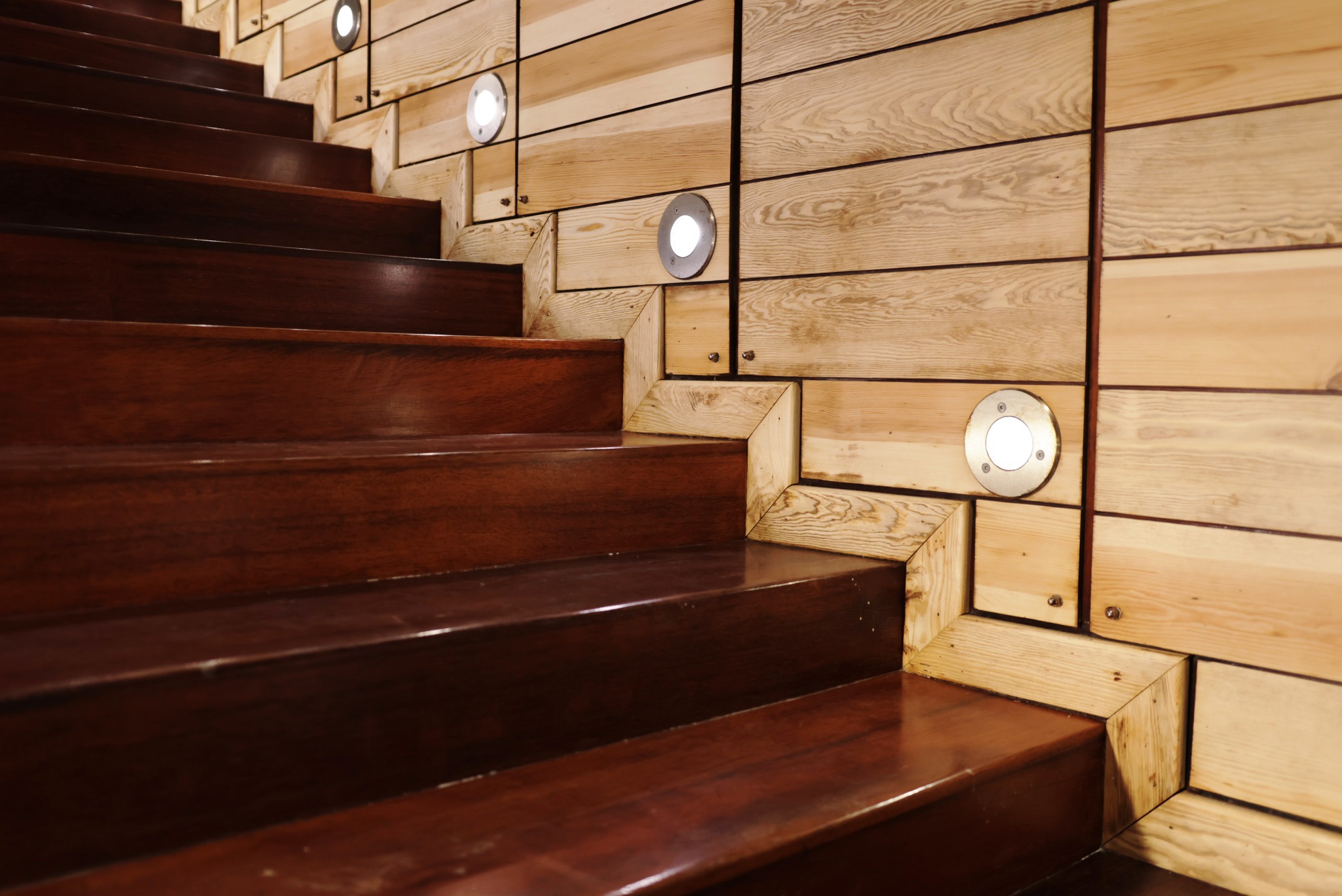 Drewniane schody z ciemnego drewna, oświetlone punktowymi lampami w ścianie z jasnego drewna. Nietypowe wykończenie ścian z drewnianych elementów.