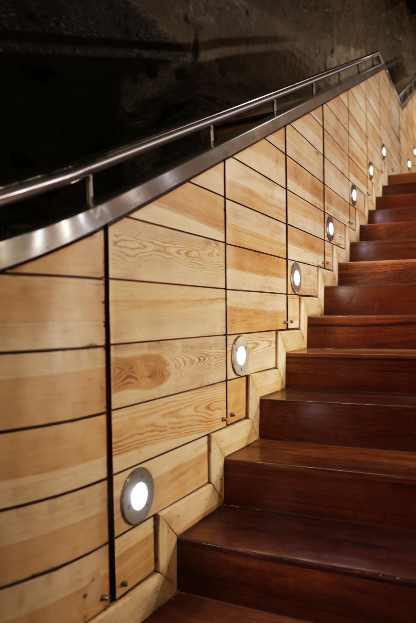 Drewniane schody z oświetleniem punktowym i metalową barierką, prowadzące w górę, z widoczną strukturą drewna i ciemnym tłem.