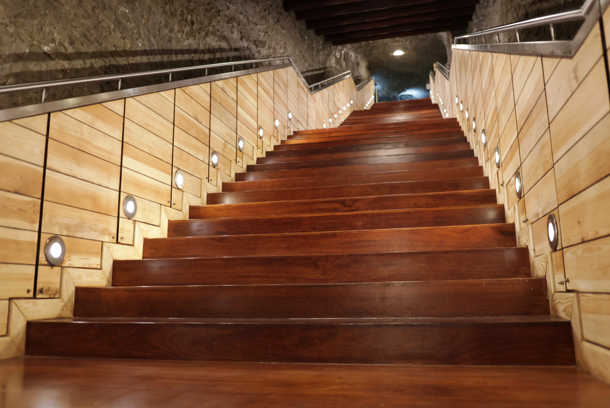Drewniane schody z oświetleniem punktowym wzdłuż ściany z jasnego drewna, prowadzące w górę do oświetlonego tunelu. Metalowa barierka po lewej stronie.