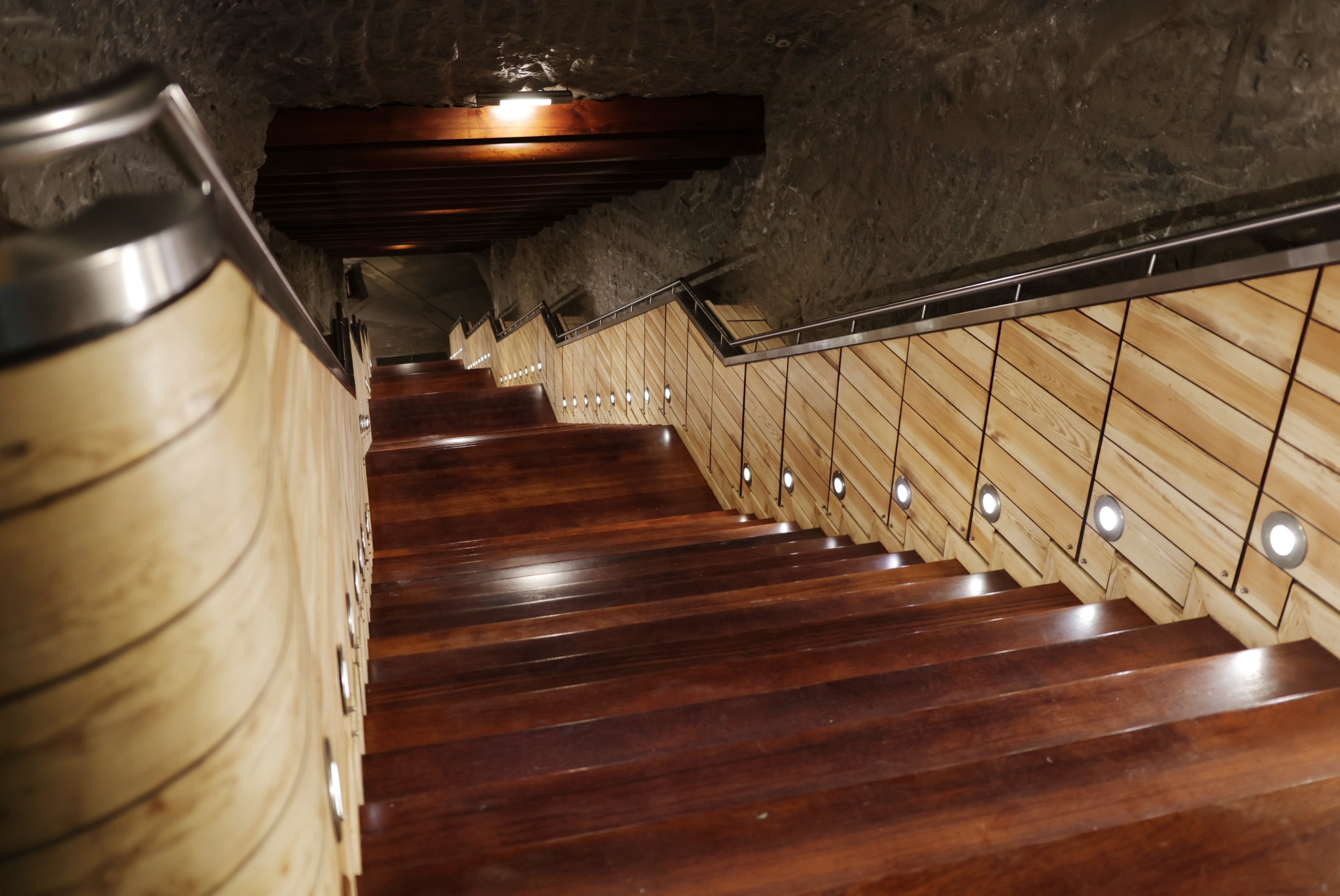 Schody z ciemnego drewna z podświetleniem LED wzdłuż jasnych, drewnianych ścianek, prowadzące w dół w kamiennym tunelu z metalową poręczą.